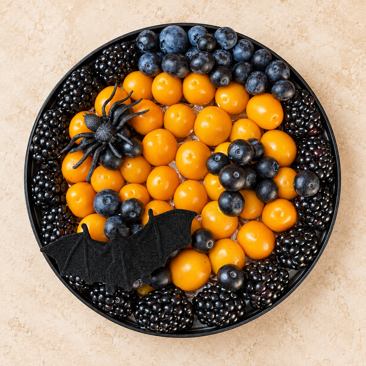A Halloween-themed fruit platter in a black round tray, featuring orange mandarin oranges, blackberries, blueberries, and black raspberries, decorated with a black plastic spider and a black felt bat on a beige stone background.