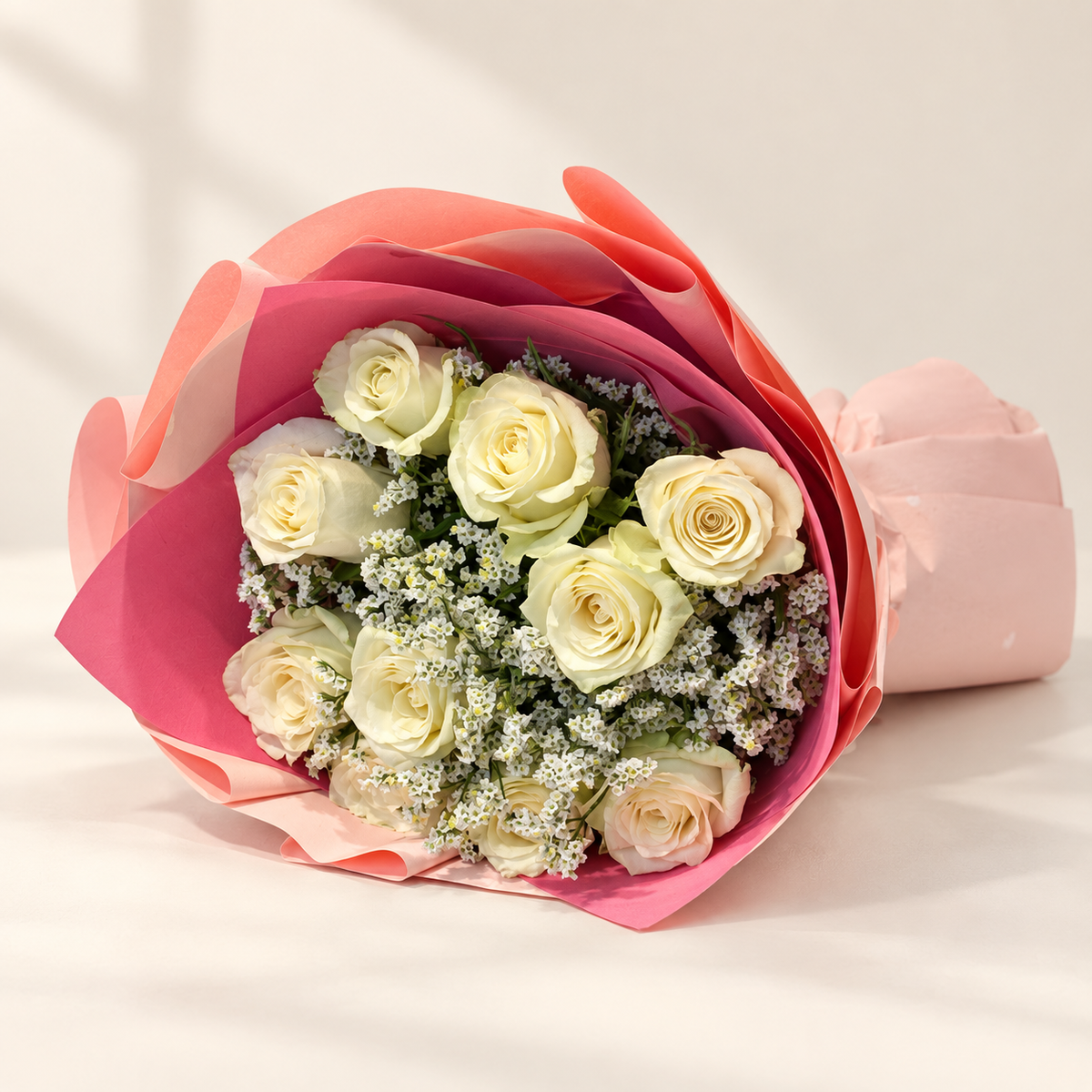 A bouquet of pale cream roses wrapped in pink and coral paper, with small white filler flowers, photographed on a clean white background.