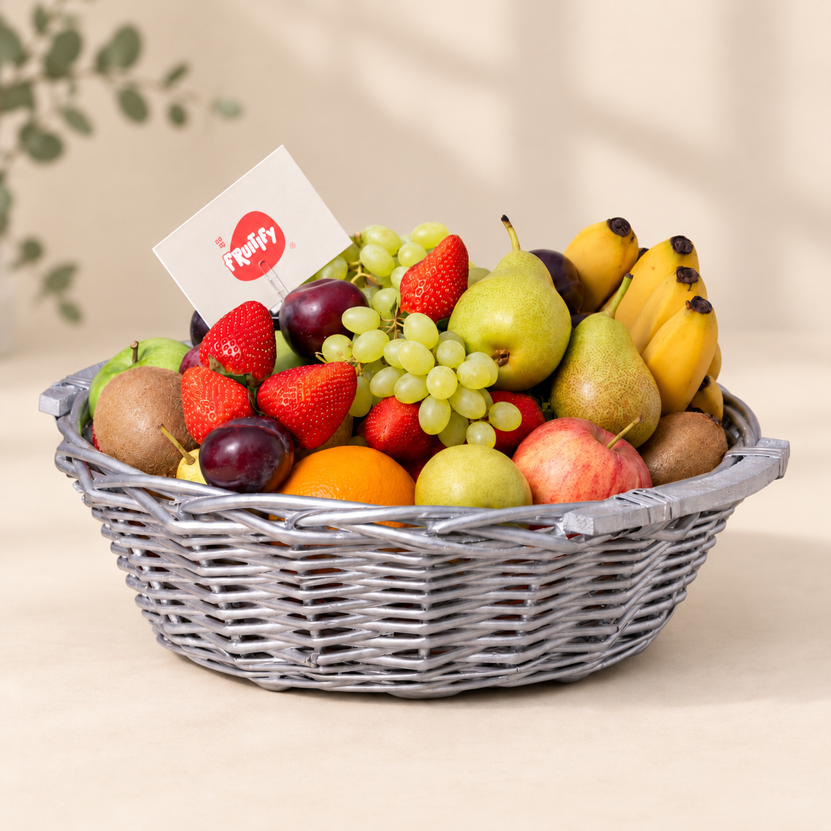 A gray wicker fruit basket filled with assorted fresh fruits including pears, bananas, grapes, strawberries, apples, kiwi, and oranges, with a small card in the corner, set against a warm beige background.
