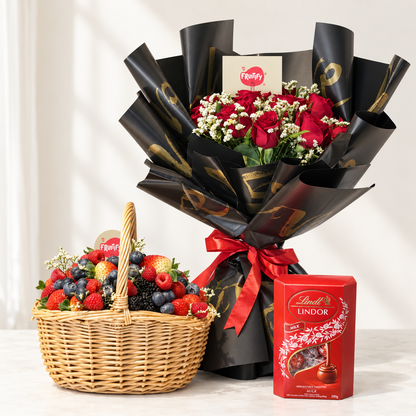 A gift set featuring a large bouquet of red roses wrapped in black paper with gold accents and a red ribbon, a wicker basket filled with fresh strawberries, blueberries, and raspberries, and a red box of Lindor Lindor chocolate.