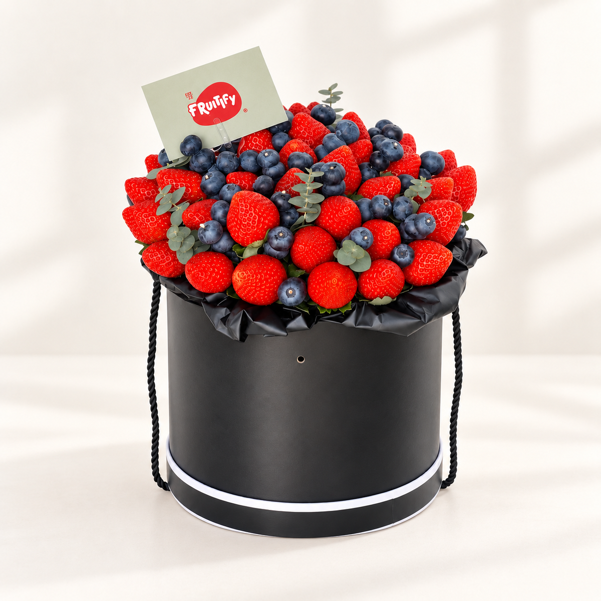 A black cylindrical gift box filled with fresh strawberries, blueberries, and small green leaves, with a small card reading 'Fronthy' placed inside. The box has black rope handles and a white stripe near the base, photographed on a clean light background.