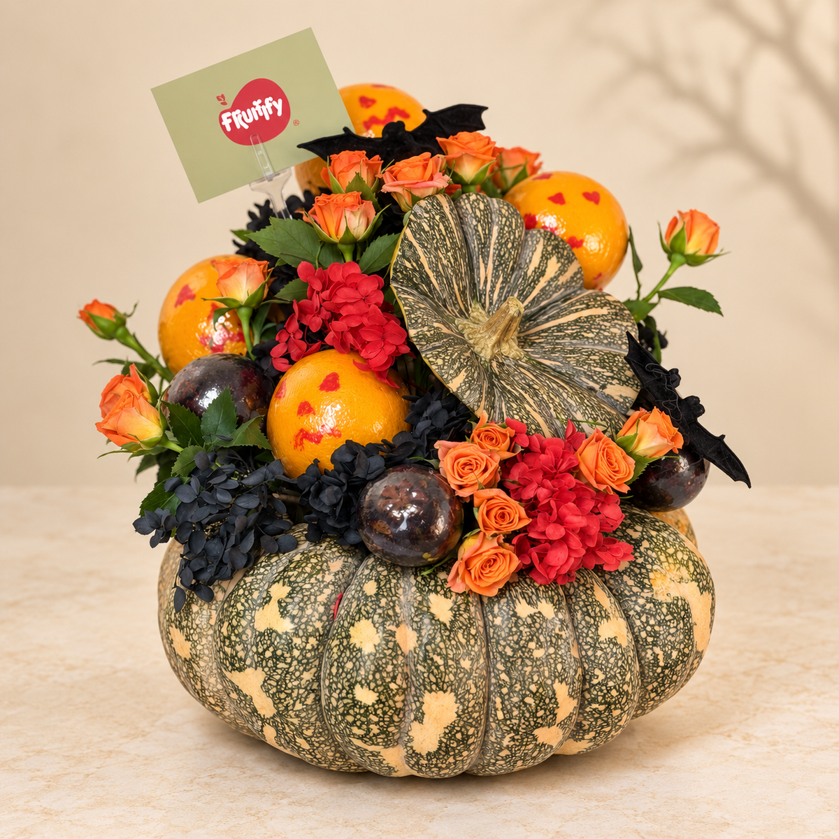 A decorative autumn floral arrangement in a speckled green and beige pumpkin-shaped planter, filled with orange marigold-like flowers, red roses, dark blue blossoms, black bat-shaped ornaments, and a striped gourd, with a small Fruffy card at the top, set on a beige surface against a soft beige background.