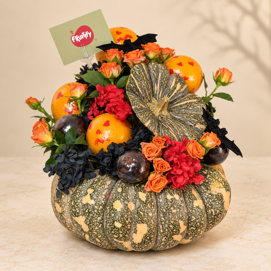 A decorative autumn floral arrangement in a speckled green and beige pumpkin-shaped planter, filled with orange marigold-like flowers, red roses, dark blue blossoms, black bat-shaped ornaments, and a striped gourd, with a small Fruffy card at the top, set on a beige surface against a soft beige background.