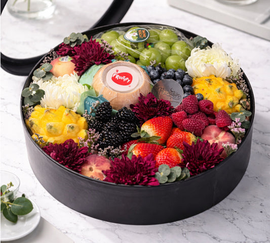 Decorative fruit and flower arrangement with a cake in a black container on a marble surface.