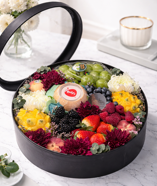 Fruit and flower arrangement in a black container on a marble surface with candles in the background.
