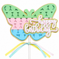 A butterfly-shaped birthday cake topper on a white stick, featuring pastel green, pink, and blue sections with a gold outline and a 'happy birthday' script overlay. Three pastel ribbons in yellow, light blue, pink, and white are tied to the bottom of the topper against a plain white background.