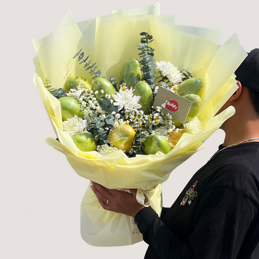 A person holding a bouquet of seasonal mangoes, with green leaves and white flowers, wrapped in yellow paper.
