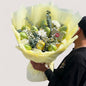 A person holding a bouquet of seasonal mangoes, with green leaves and white flowers, wrapped in yellow paper.