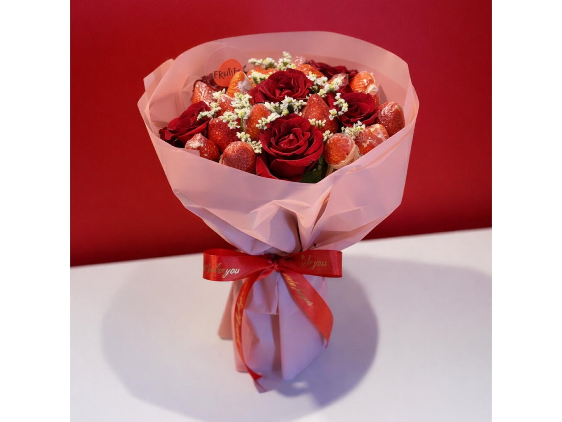 Bouquet of red roses and strawberries wrapped in pink paper with a red ribbon on a white surface.